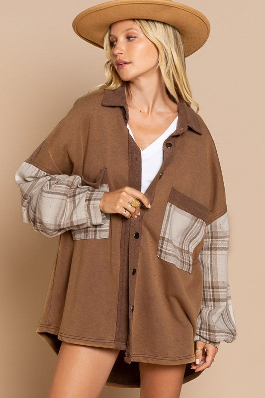 A person wearing the Long Sleeve With Plaid Detail Sleeve Shacket in brown, featuring plaid details on the sleeves and a matching pocket, paired with a white top underneath and a wide-brimmed hat—perfect for transitional weather.