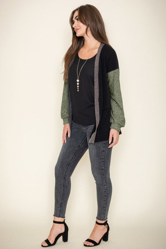 A woman is smiling and touching her hair while wearing the Color Block Knit Cardigan, featuring shades of black, grey, and beige. She has paired it with a black top and grey jeans. The cardigan is made from 95% rayon and is proudly manufactured in the United States.