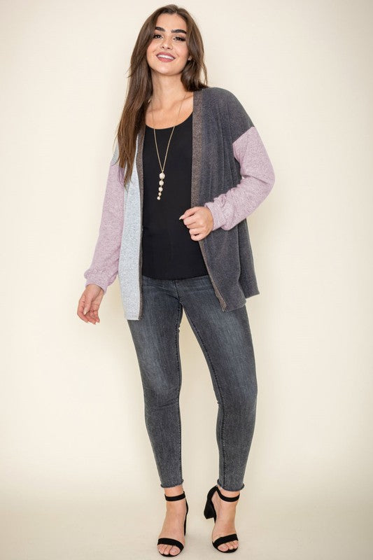 A woman is smiling and touching her hair while wearing the Color Block Knit Cardigan, featuring shades of black, grey, and beige. She has paired it with a black top and grey jeans. The cardigan is made from 95% rayon and is proudly manufactured in the United States.