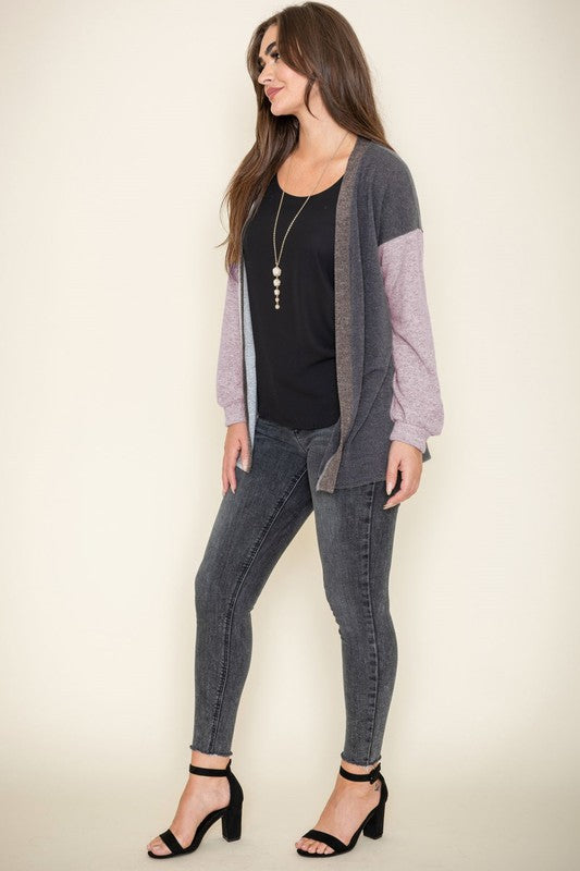 A woman is smiling and touching her hair while wearing the Color Block Knit Cardigan, featuring shades of black, grey, and beige. She has paired it with a black top and grey jeans. The cardigan is made from 95% rayon and is proudly manufactured in the United States.