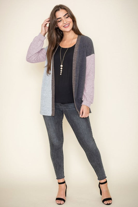 A woman is smiling and touching her hair while wearing the Color Block Knit Cardigan, featuring shades of black, grey, and beige. She has paired it with a black top and grey jeans. The cardigan is made from 95% rayon and is proudly manufactured in the United States.