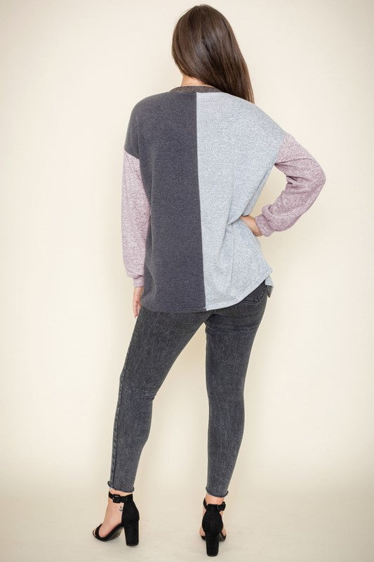 A woman is smiling and touching her hair while wearing the Color Block Knit Cardigan, featuring shades of black, grey, and beige. She has paired it with a black top and grey jeans. The cardigan is made from 95% rayon and is proudly manufactured in the United States.