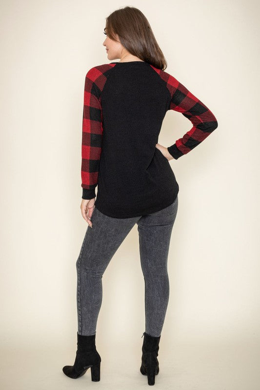 A woman with curly hair, wearing a Plus Checker Sleeve Sequin Pocket Tunic featuring dark grey fabric and red plaid checker sleeves, stands against a white background. Made in the United States, she has her hand in her pocket and is smiling.