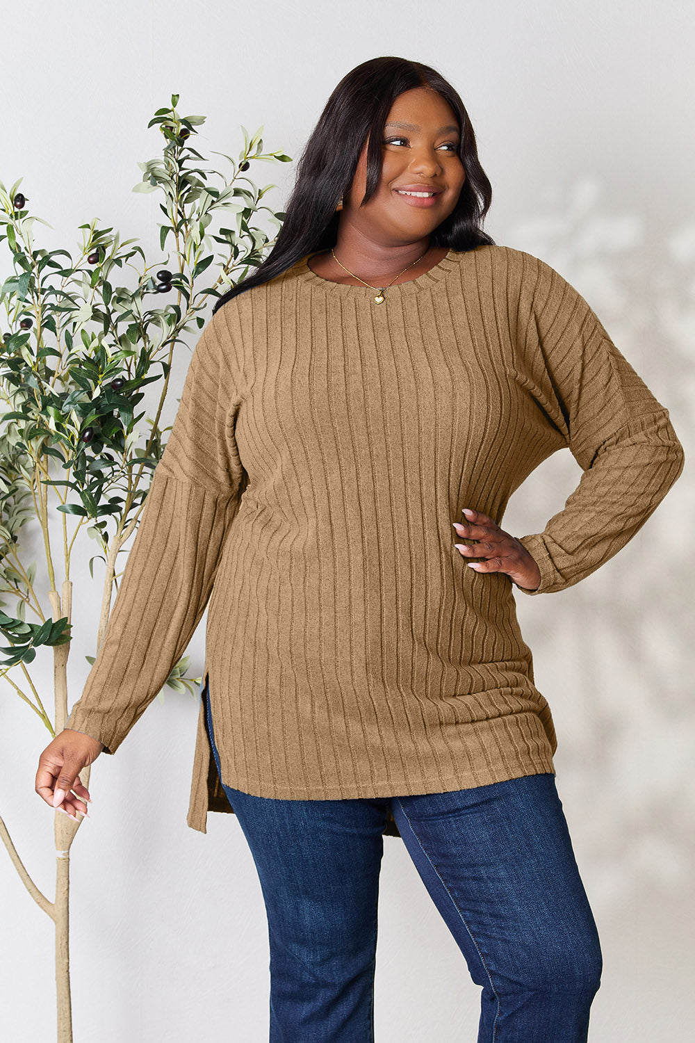 A person with shoulder-length hair smiles while wearing a beige ribbed sweater called the Basic Bae Full Size Ribbed Round Neck Long Sleeve Slit Top, along with matching beige pants, showcasing a basic style. They stand next to a green plant.