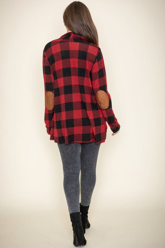 A woman stands against a plain background, wearing the Checkered Elbow Patch Cardigan with stylish elbow patches in red and black, paired with a black top, gray jeans, and black ankle boots. The ensemble captures a timeless look that's effortlessly chic.