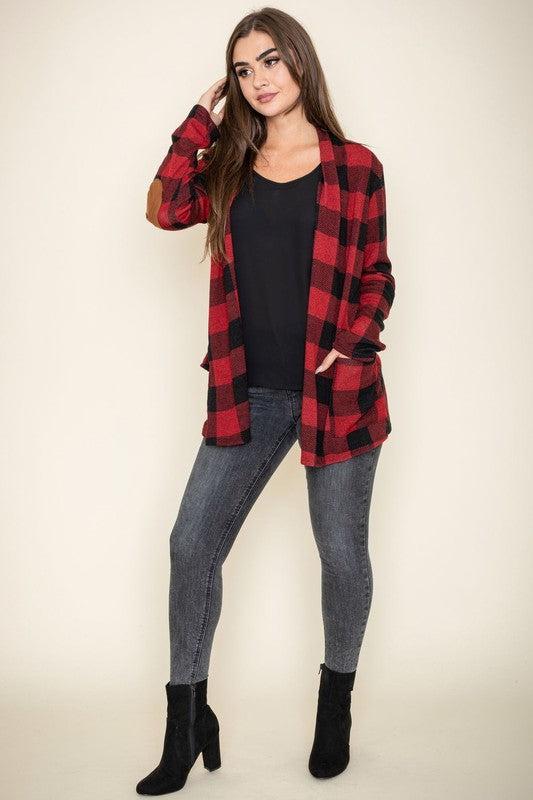 A woman stands against a plain background, wearing the Checkered Elbow Patch Cardigan with stylish elbow patches in red and black, paired with a black top, gray jeans, and black ankle boots. The ensemble captures a timeless look that's effortlessly chic.