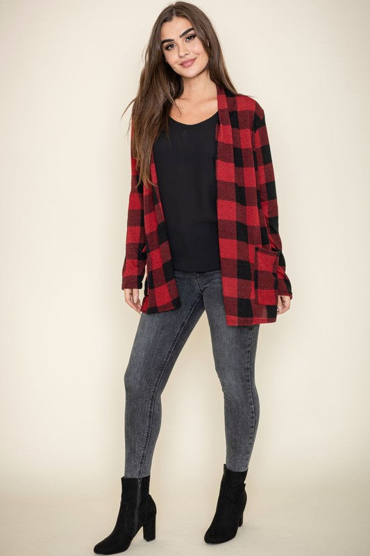 A woman stands against a plain background, wearing the Checkered Elbow Patch Cardigan with stylish elbow patches in red and black, paired with a black top, gray jeans, and black ankle boots. The ensemble captures a timeless look that's effortlessly chic.