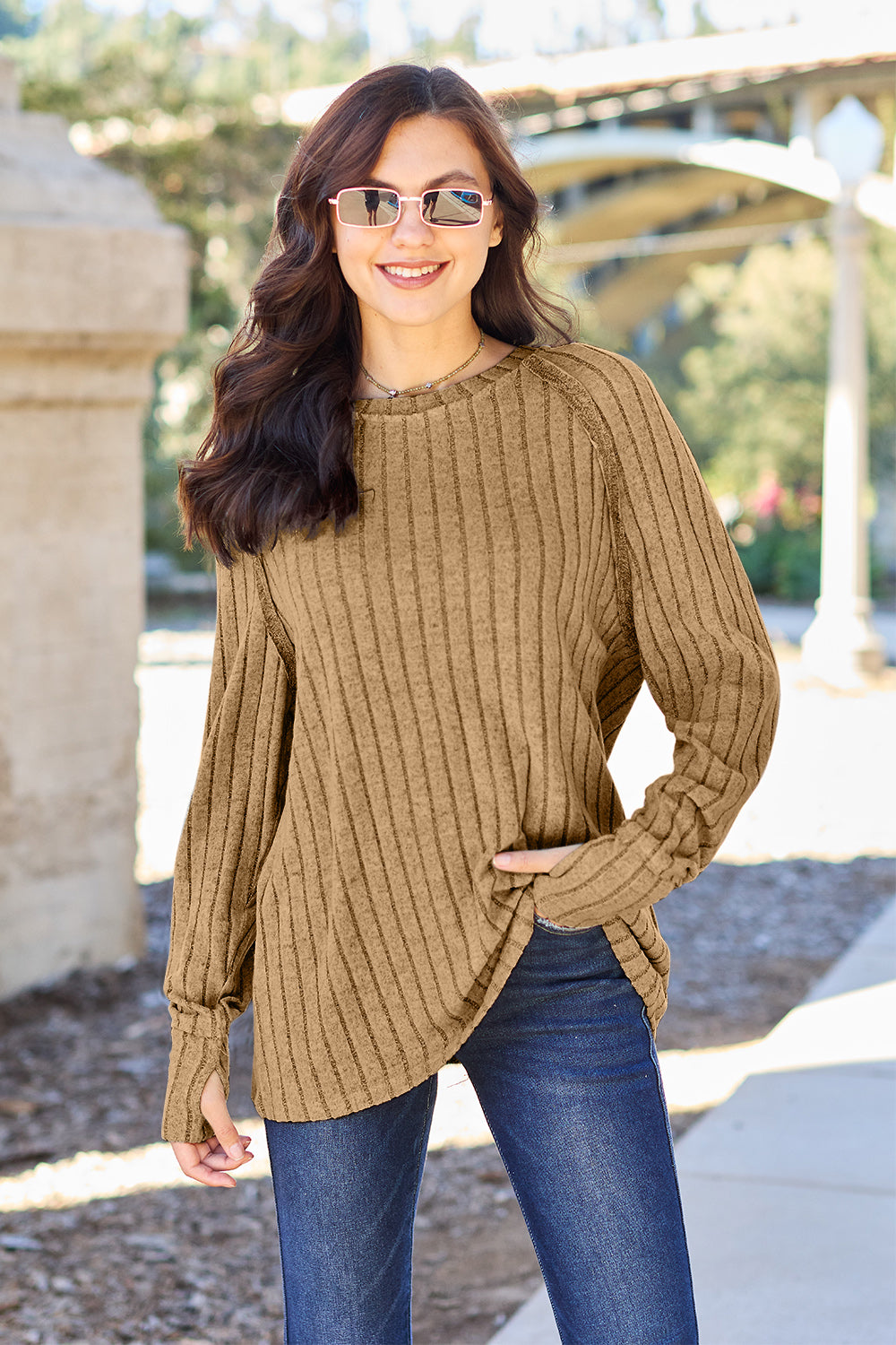 A person with long dark hair and sunglasses stands outdoors, wearing the Basic Bae Full Size Ribbed Round Neck Long Sleeve Knit Top in blue and a pair of jeans. The basic style outfit blends seamlessly with the background of trees and a bridge.