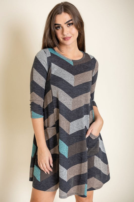 A person stands against a plain background, showcasing the Plus Chevron A-Line Dress in grey, taupe, and teal. The dress features three-quarter sleeves and is crafted from a polyester spandex blend, effortlessly combining comfort and style.