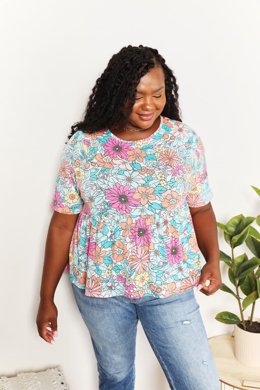 A person wearing a Double Take Floral Round Neck Babydoll Top and jeans stands with one hand in their pocket, with a plant in the background.