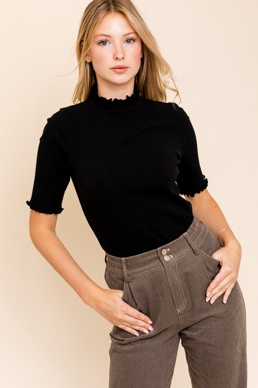 A person in a rust-colored Short Sleeve Mock Neck Merrow Edge Top holds a beige hat. They are also wearing a pleated skirt.