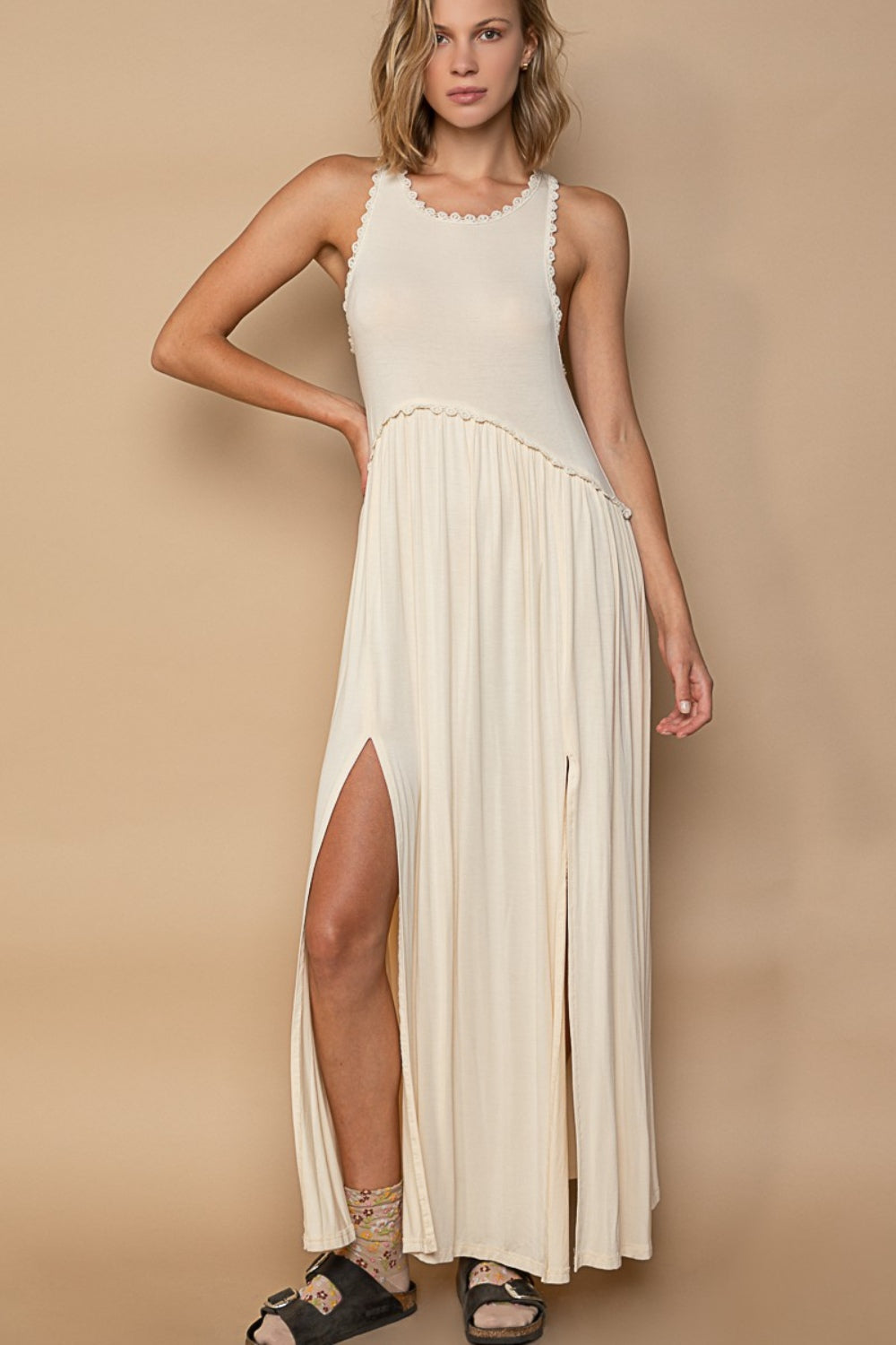 A woman wearing a POL Sleeveless Back Zipper Front Slit Maxi Dress in cream stands against a beige background. She has one hand on her hip and is wearing black sandals and patterned socks.