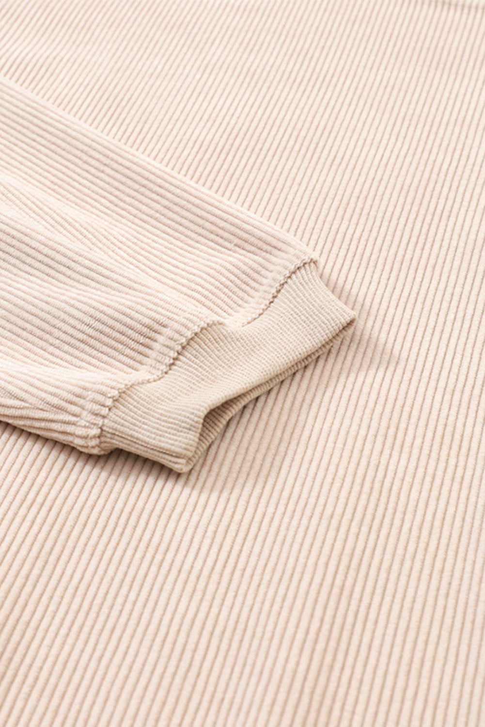 A woman stands outdoors, looking down while touching her hair with one hand. She is wearing an Apricot Ribbed Corduroy Oversized Sweatshirt and white pants, exuding an effortlessly chic and fashionable look.