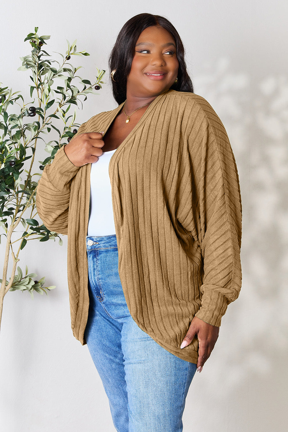 A person with wavy hair, wearing the Basic Bae Full Size Ribbed Cocoon Cardigan over a white top and beige pants, stands in front of a light-colored background with a leafy plant, showcasing an effortlessly basic style.