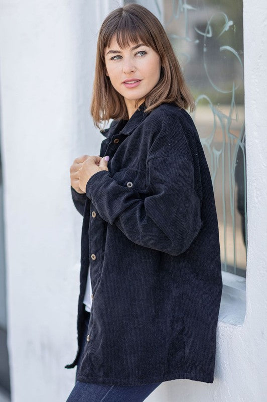 The person is smiling and glancing sideways, dressed in a light gray button-up shirt layered over a white top, exuding an effortlessly stylish vibe. Their outfit mirrors the relaxed elegance of the Corduroy Jacket while delivering a breezier feel for casual comfort.