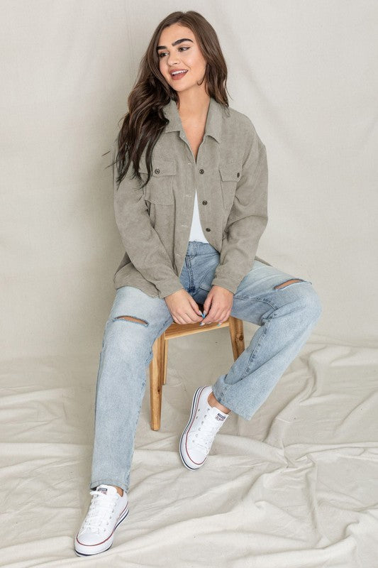 The person is smiling and glancing sideways, dressed in a light gray button-up shirt layered over a white top, exuding an effortlessly stylish vibe. Their outfit mirrors the relaxed elegance of the Corduroy Jacket while delivering a breezier feel for casual comfort.