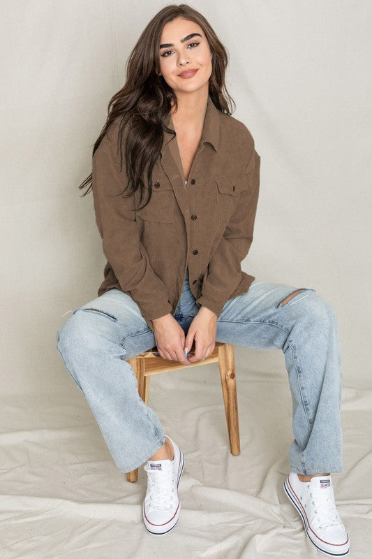 The person is smiling and glancing sideways, dressed in a light gray button-up shirt layered over a white top, exuding an effortlessly stylish vibe. Their outfit mirrors the relaxed elegance of the Corduroy Jacket while delivering a breezier feel for casual comfort.