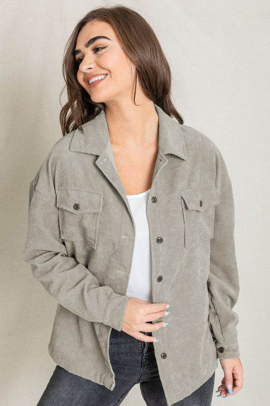 The person is smiling and glancing sideways, dressed in a light gray button-up shirt layered over a white top, exuding an effortlessly stylish vibe. Their outfit mirrors the relaxed elegance of the Corduroy Jacket while delivering a breezier feel for casual comfort.