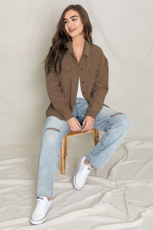 The person is smiling and glancing sideways, dressed in a light gray button-up shirt layered over a white top, exuding an effortlessly stylish vibe. Their outfit mirrors the relaxed elegance of the Corduroy Jacket while delivering a breezier feel for casual comfort.