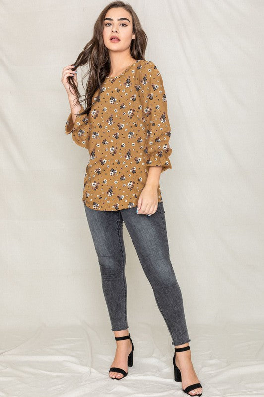 A woman with long brown hair is wearing the Floral Ruffle Sleeve Tunic in mustard yellow, made of 100% polyester, along with dark jeans, standing against a light-colored background.