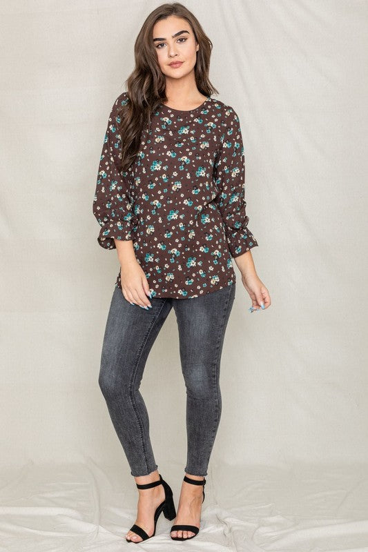 A woman with long brown hair is wearing the Floral Ruffle Sleeve Tunic in mustard yellow, made of 100% polyester, along with dark jeans, standing against a light-colored background.