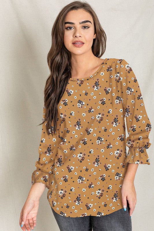 A woman with long brown hair is wearing the Floral Ruffle Sleeve Tunic in mustard yellow, made of 100% polyester, along with dark jeans, standing against a light-colored background.