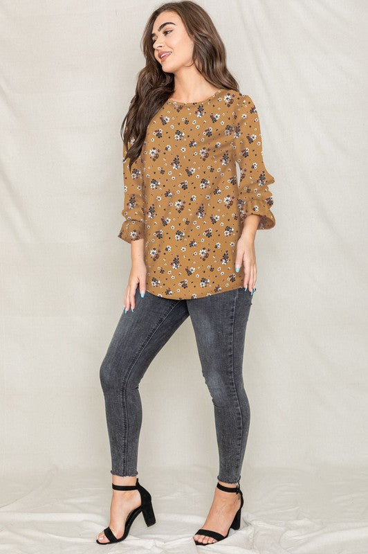 A woman with long brown hair is wearing the Floral Ruffle Sleeve Tunic in mustard yellow, made of 100% polyester, along with dark jeans, standing against a light-colored background.
