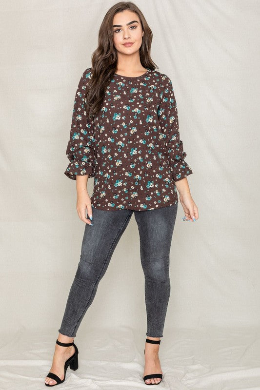 A woman with long brown hair is wearing the Floral Ruffle Sleeve Tunic in mustard yellow, made of 100% polyester, along with dark jeans, standing against a light-colored background.