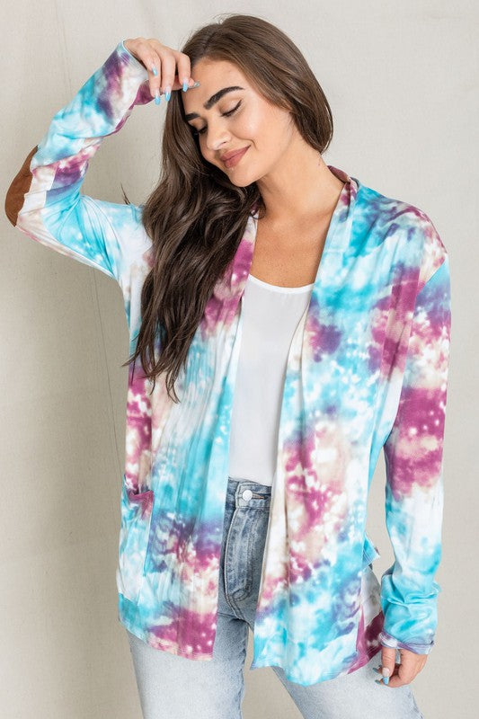 A woman poses for a picture, highlighting her stylish Tie Dye Elbow Patch Open Cardigan. The vibrant colors and unique design emphasize the craftsmanship of this piece, proudly made in the United States.