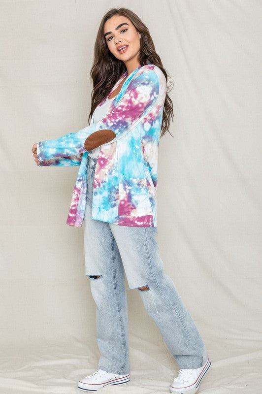 A woman poses for a picture, highlighting her stylish Tie Dye Elbow Patch Open Cardigan. The vibrant colors and unique design emphasize the craftsmanship of this piece, proudly made in the United States.