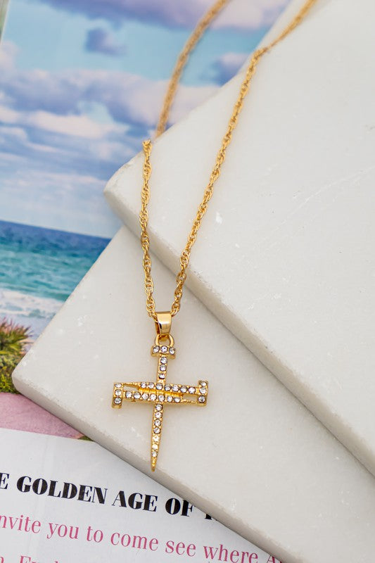 A Nail shape cross pendant necklace with a rope chain, featuring small gemstones, rests elegantly on a marble surface. In the background, an open magazine reveals a tranquil beach scene.