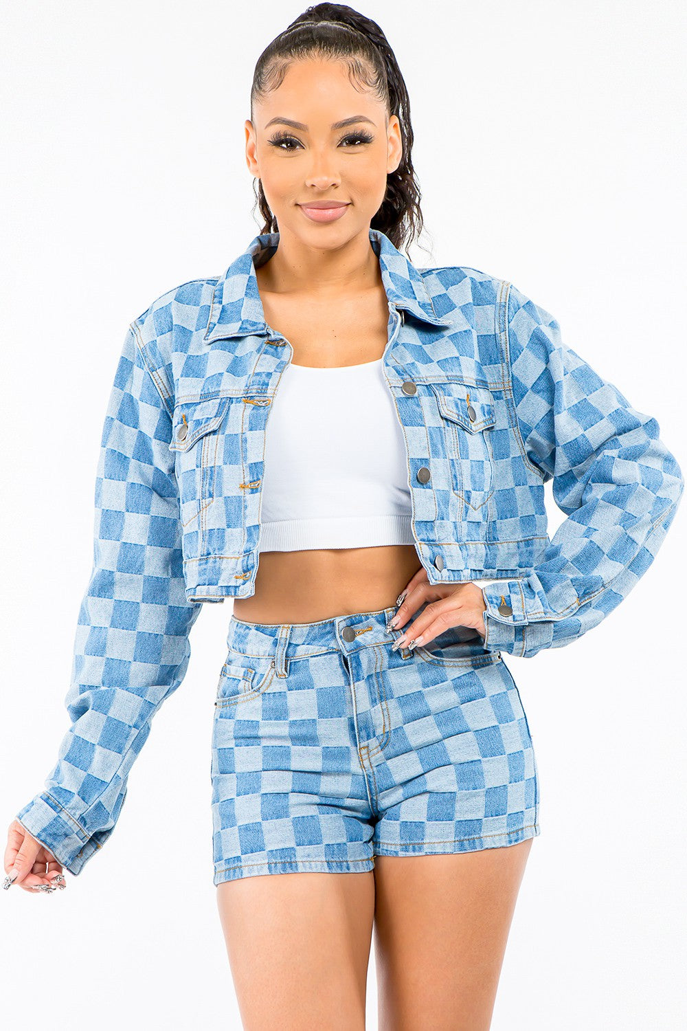 A woman stands against a white background wearing an American Bazi Checkered Long Sleeve Cropped Denim Jacket, paired with a white crop top and matching blue checkered denim shorts. Her hair is tied back in a high ponytail.