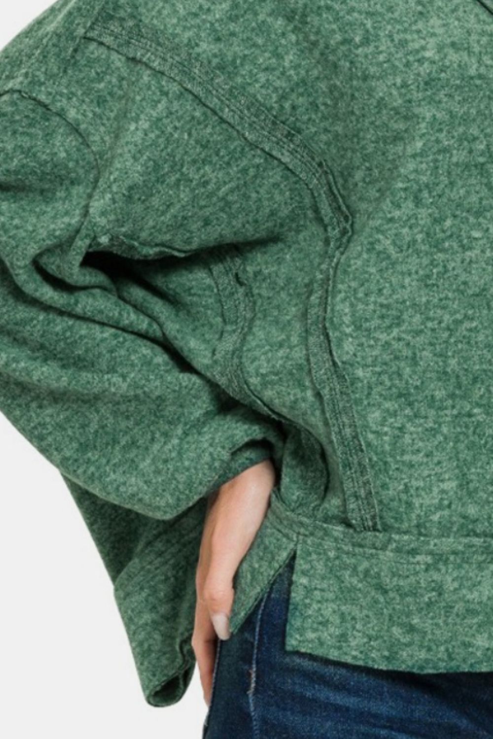 A person wearing a Zenana Brushed Hacci Exposed Seam Hoodie in green, complemented by jeans, poses confidently with a hand on their hip.