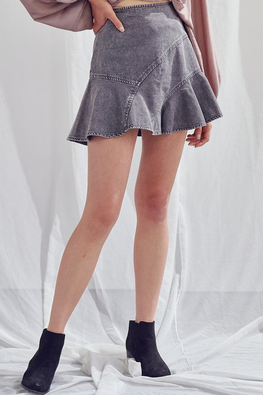 Person wearing a light-colored, acid wash A-Line skirt at mini length, paired with a long-sleeve black top and black ankle boots stands against a white background.