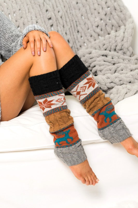 A person wearing Nordic Snowflake Leg Warmers and a grey sweater sits on a white couch with a grey blanket draped over it.