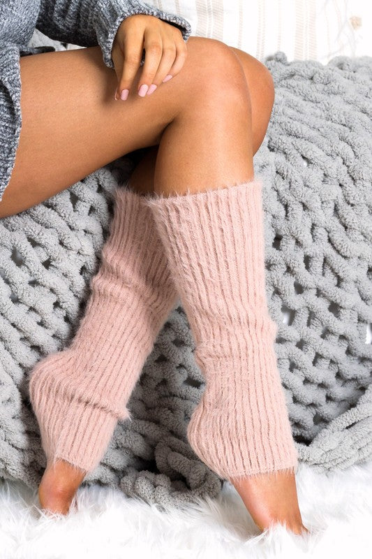 Person seated on a gray knitted blanket, wearing pink, fuzzy knee-high socks and a gray sweater, with Eyelash Leg Warmers adding an extra touch of comfort.