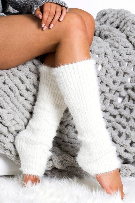 A person wearing Eyelash Leg Warmers made from buttery soft-spun acrylic sits on a gray knitted blanket, their hands resting on their knees.