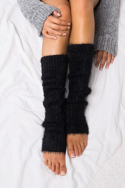 A person is sitting on a gray textured blanket, wearing a gray sweater and Eyelash Leg Warmers in pink, with their legs crossed and hands resting on their knees.
