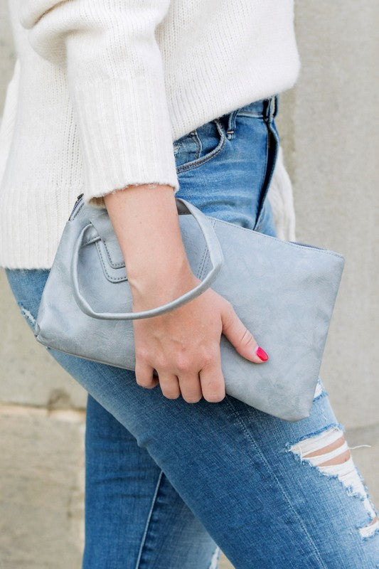 Person in a light sweater and ripped jeans holding a compact size, tan Wristlet Mini Clutch made of vegan PU leather.