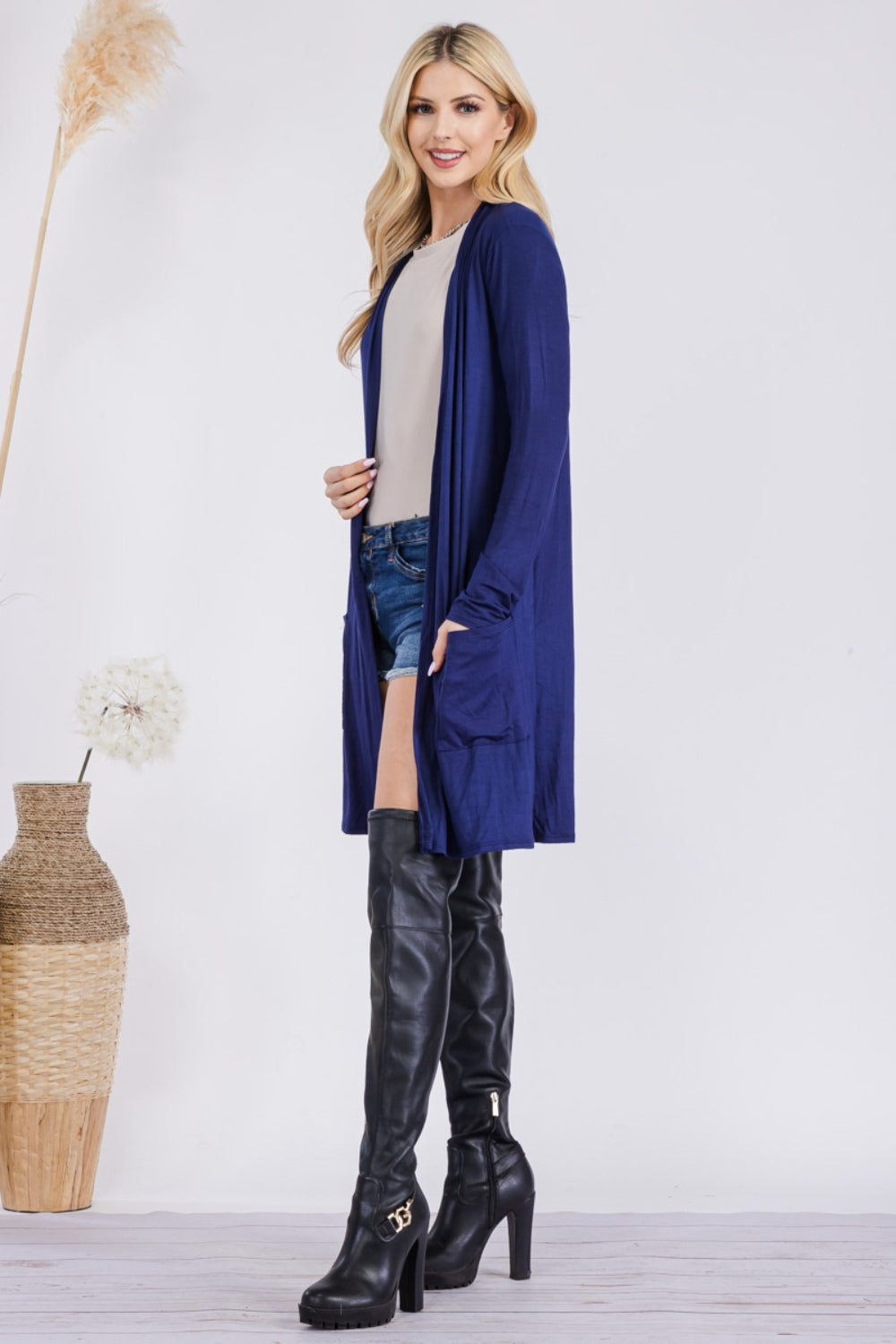 A woman stands indoors wearing the Celeste Full Size Open Front Cardigan with Pockets, a long blue USA-made cardigan. She pairs it with a beige top, denim shorts, and black thigh-high boots. She is smiling and looking to her left, with tall vases of dried plants beside her.