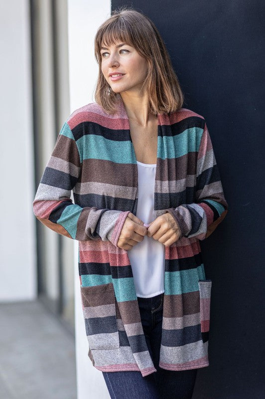 A person wearing the Plus Suede Elbow Patch Open Cardigan with Pockets, characterized by its multi-colored stripes, suede elbow patches, and dual front pockets, stands against a plain background over a white shirt and jeans.