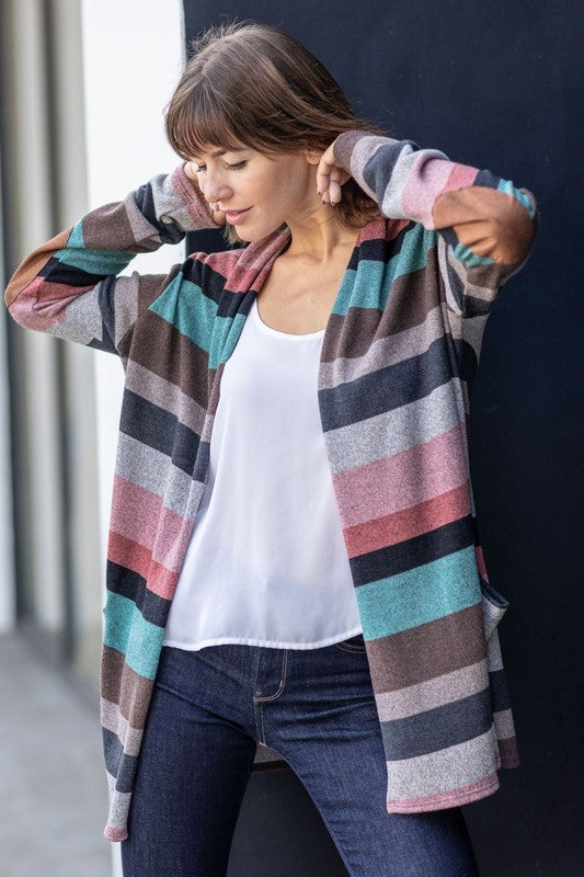 A person wearing the Plus Suede Elbow Patch Open Cardigan with Pockets, characterized by its multi-colored stripes, suede elbow patches, and dual front pockets, stands against a plain background over a white shirt and jeans.