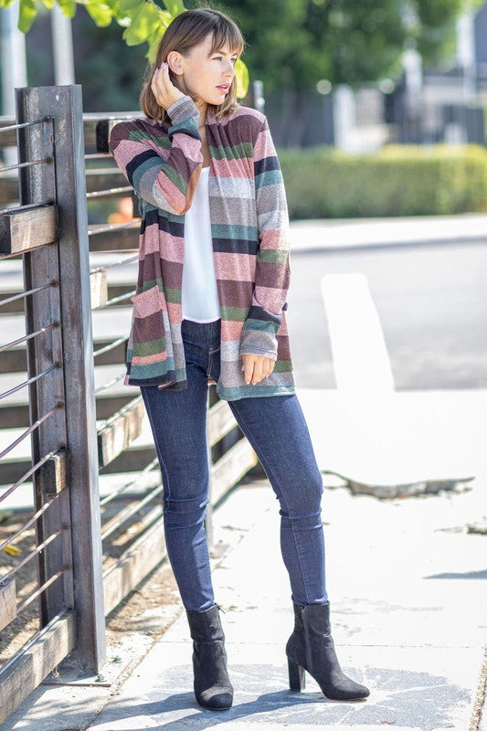 A person wearing the Plus Suede Elbow Patch Open Cardigan with Pockets, characterized by its multi-colored stripes, suede elbow patches, and dual front pockets, stands against a plain background over a white shirt and jeans.