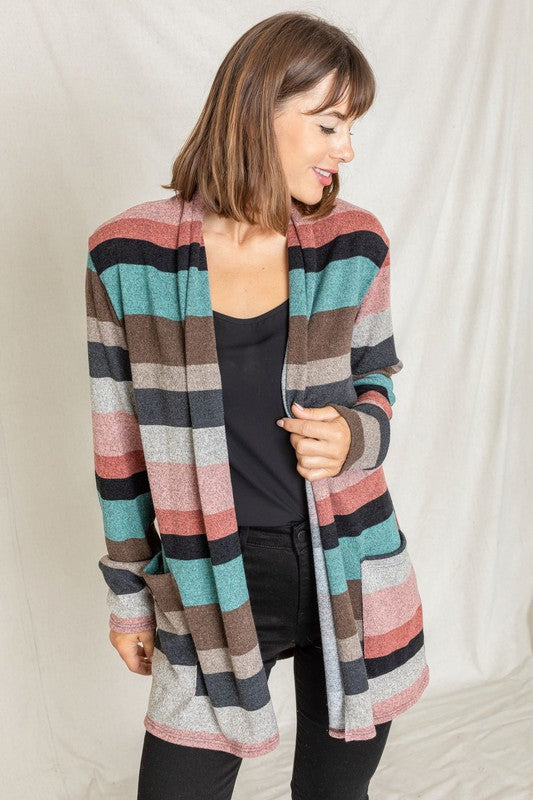 A person wearing the Plus Suede Elbow Patch Open Cardigan with Pockets, characterized by its multi-colored stripes, suede elbow patches, and dual front pockets, stands against a plain background over a white shirt and jeans.