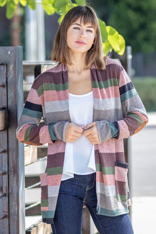 A person wearing the Plus Suede Elbow Patch Open Cardigan with Pockets, characterized by its multi-colored stripes, suede elbow patches, and dual front pockets, stands against a plain background over a white shirt and jeans.