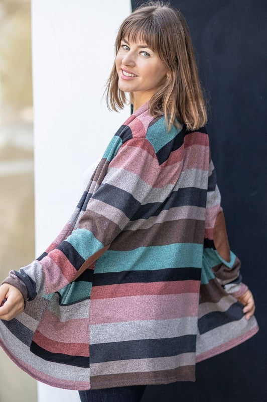 A person wearing the Plus Suede Elbow Patch Open Cardigan with Pockets, characterized by its multi-colored stripes, suede elbow patches, and dual front pockets, stands against a plain background over a white shirt and jeans.