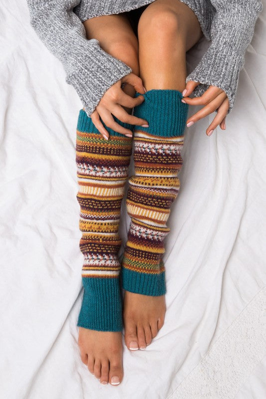 A person wearing a grey sweater and the Short Fairisle Legwarmer sits on a white bed, legs extended and hands resting gently on the soft spun acrylic.