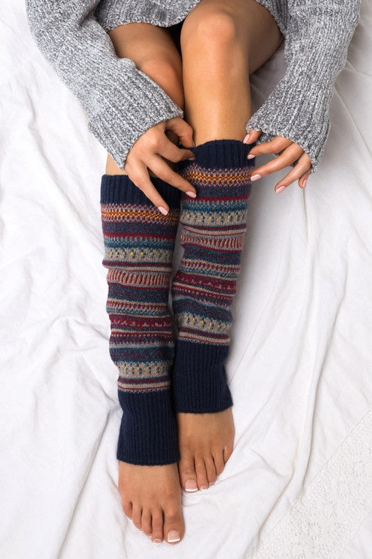 A person wearing a grey sweater and the Short Fairisle Legwarmer sits on a white bed, legs extended and hands resting gently on the soft spun acrylic.