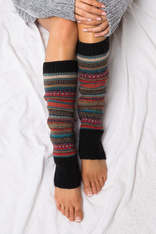 A person wearing a grey sweater and the Short Fairisle Legwarmer sits on a white bed, legs extended and hands resting gently on the soft spun acrylic.