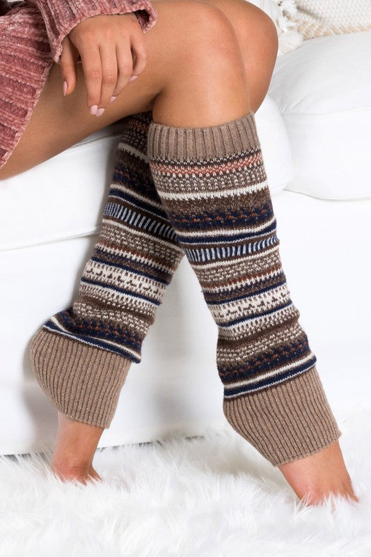 A person wearing a grey sweater and the Short Fairisle Legwarmer sits on a white bed, legs extended and hands resting gently on the soft spun acrylic.